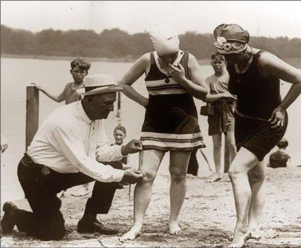 Foto antiga, da década de 40, de um homem com fita métrica medindo o comprimento da saia de mulher na praia.